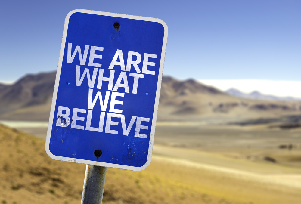 Blue sign with white letters on the side of desert road that says, "We are what we believe."
